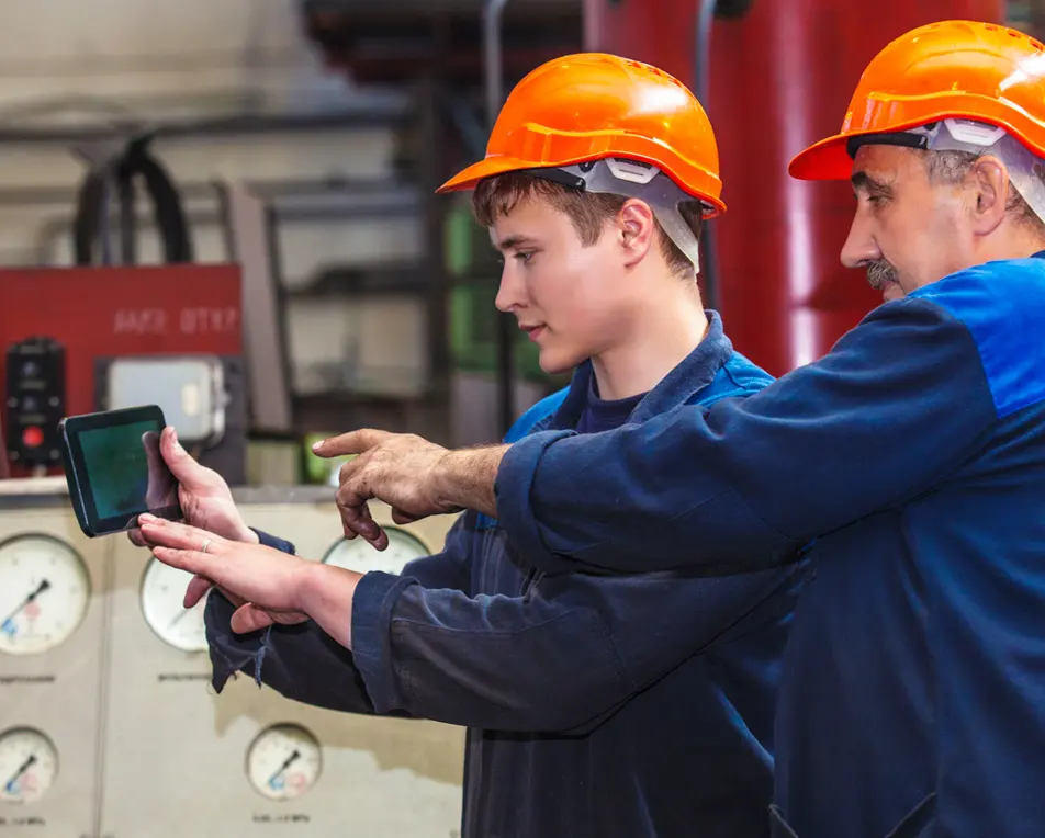 Twee personen met oranje helmen en blauwe werkkleding overleggen bij een tablet in een industriële omgeving met drukmeters op de achtergrond.