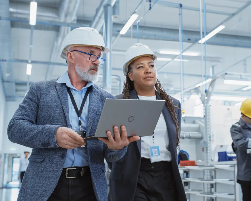 Twee personen met witte helmen en zakelijke kleding lopen door een industriële omgeving; één van hen toont iets op een laptop.