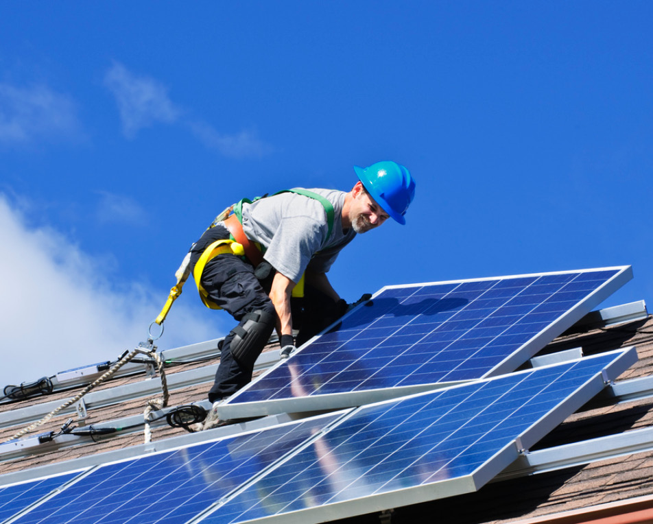 Een monteur met blauwe helm en veiligheidsharnas installeert zonnepanelen op een dak onder een heldere blauwe lucht.