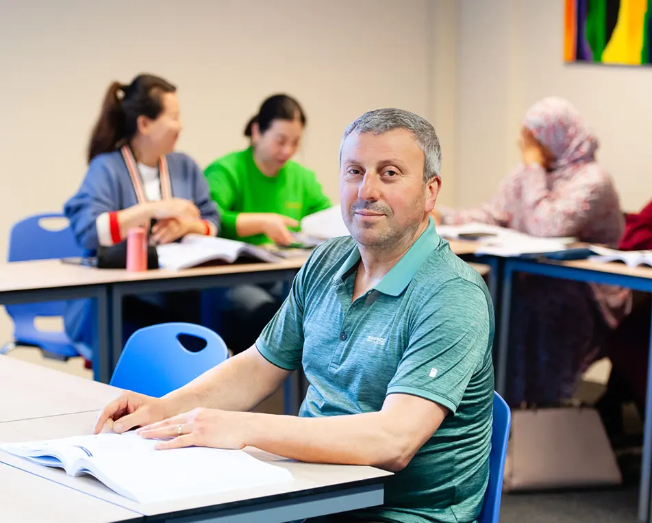 Vier personen zitten in een klaslokaal aan bureaus met boeken; op de voorgrond een oudere persoon in een groene polo, op de achtergrond drie anderen in gesprek of lezend.