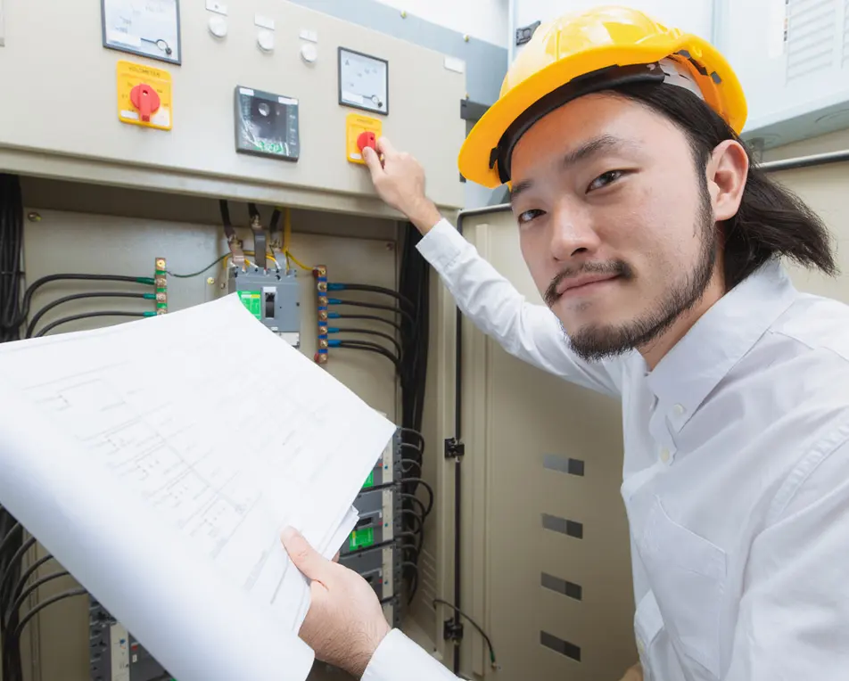 Persoon met gele bouwhelm en wit shirt bedient een elektrisch bedieningspaneel terwijl hij een schema op papier vasthoudt.