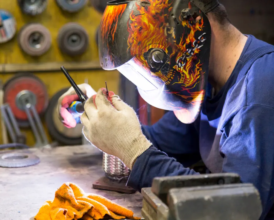 Persoon met lashelm met vlammenmotief last een metalen object aan een werkbank, met gereedschap en slijpschijven op de achtergrond.