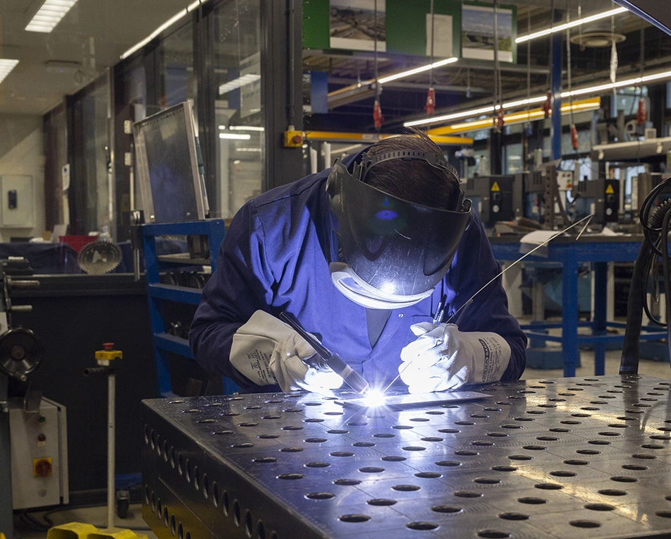 Persoon met lashelm last een metalen object aan een werkbank, met gereedschap en slijpschijven op de achtergrond.