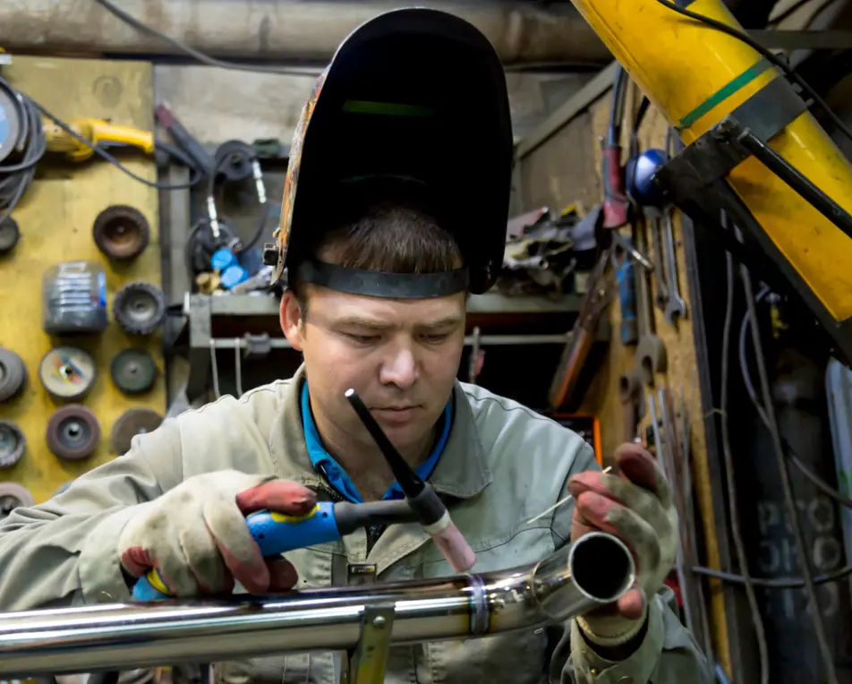 Een persoon met lashelm en handschoenen last aan een metalen pijp in een werkplaats met gereedschap aan de muur.