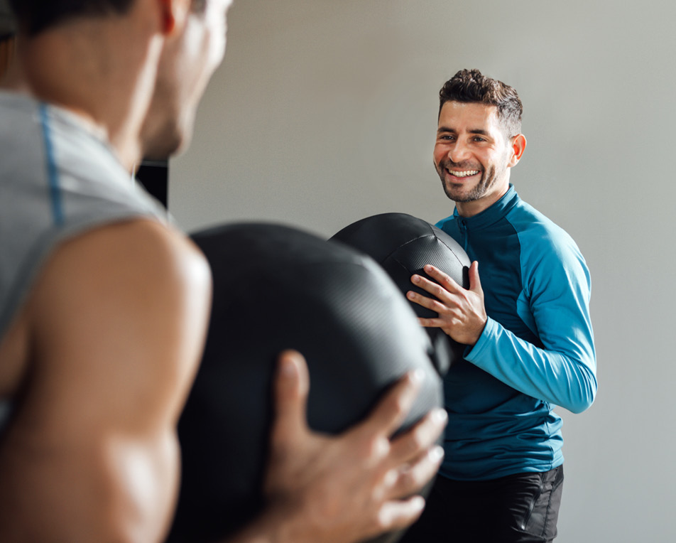 Twee personen doen een fitnessoefening met grote zwarte oefenballen; één draagt een mouwloos shirt, de ander een blauw sportshirt met lange mouwen.