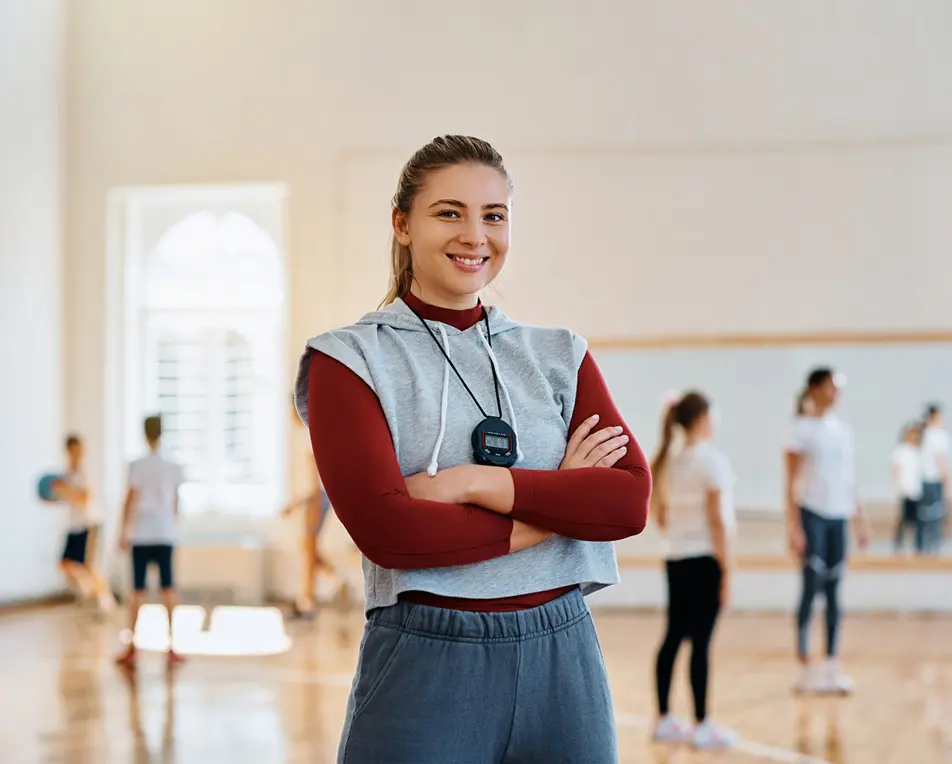 Persoon in een gymzaal met grijze mouwloze hoodie en stopwatch om de nek, terwijl anderen op de achtergrond bezig zijn met sportactiviteiten.