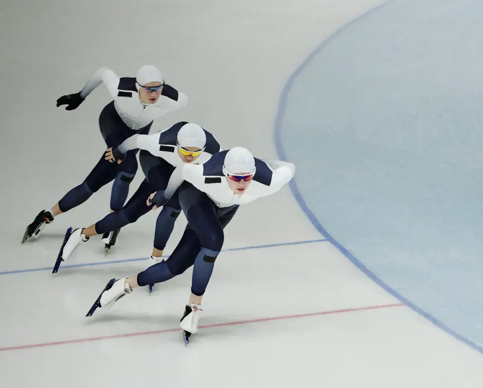 Drie schaatsers in wedstrijduniformen rijden dicht achter elkaar op een ijsbaan, met duidelijke baanaanduidingen op het ijs