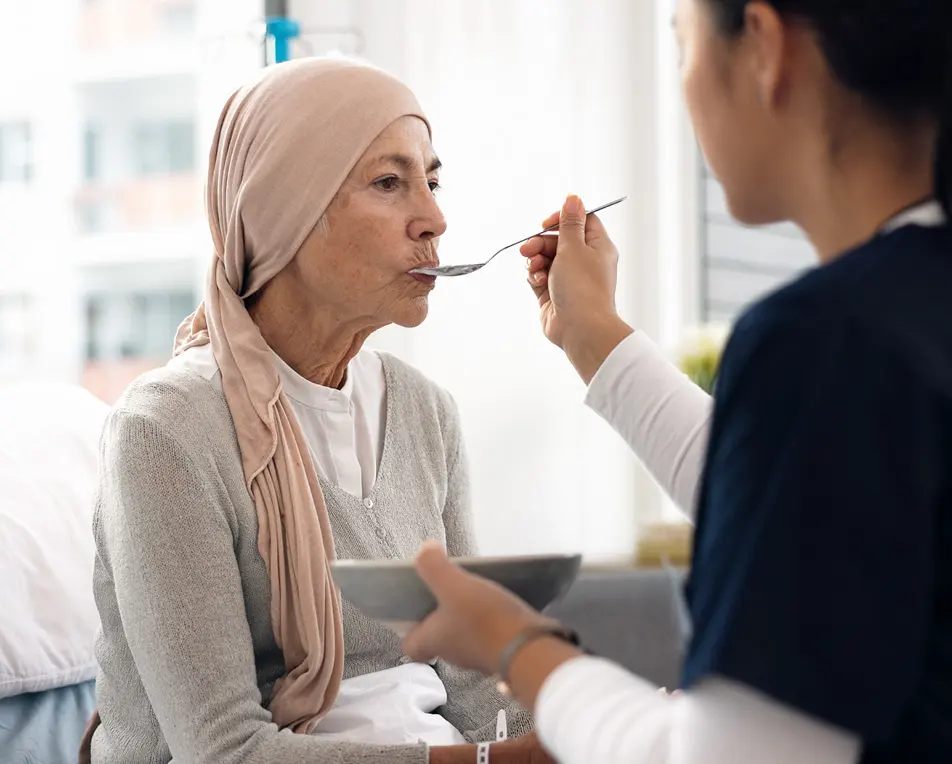 Vrouw met hoofddoek zit rechtop in bed. Andere vrouw helpt haar met eten.