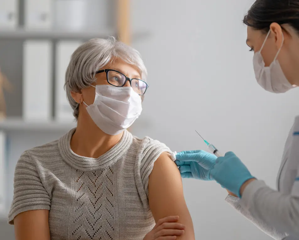Vrouw met mondkap krijgt een prik, rechts daarnaast staat een verpleegkundige met een spuit in haar hand.