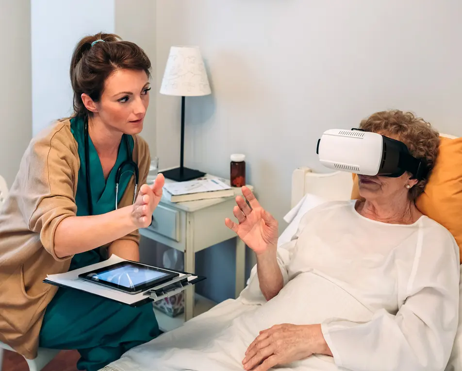 Oudere vrouw ligt in bed met een vr-bril op, daarnaast zit een vrouw in zorgkleding met een ipad.