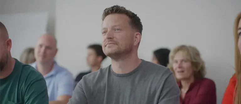 Man kijkt tevreden. Met een groep mensen in een klaslokaal op de achtergrond.