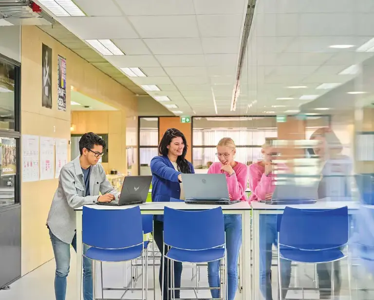 Drie personen staat rond een tafel met blauwe stoelen en laptops.
