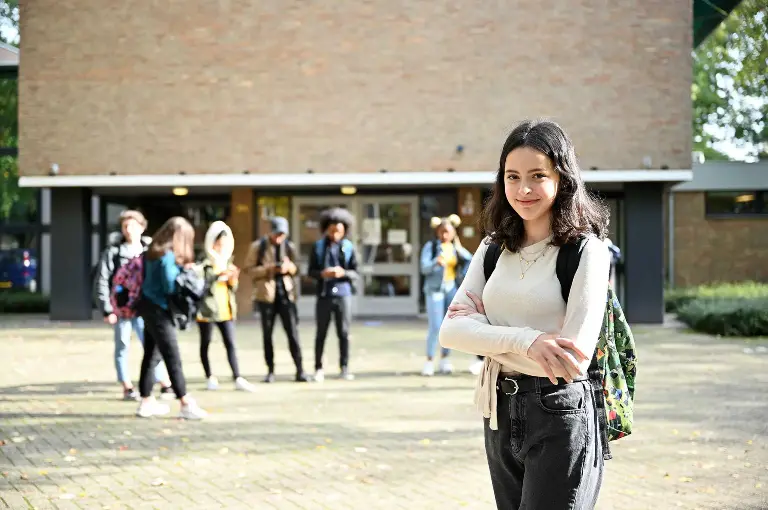 Leerling staat in beige shirt met rugzak, op de achtergrond zijn andere leerlingen en het schoolplein.