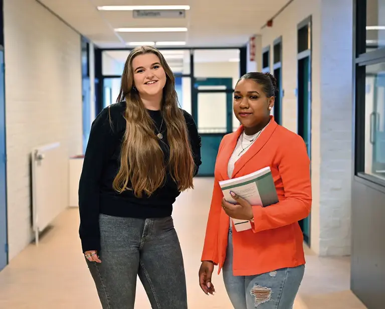 Twee vrouwen in een gang, een draagt een rode blazer en heeft een boek vast en de ander draagt een zwarte blazer.