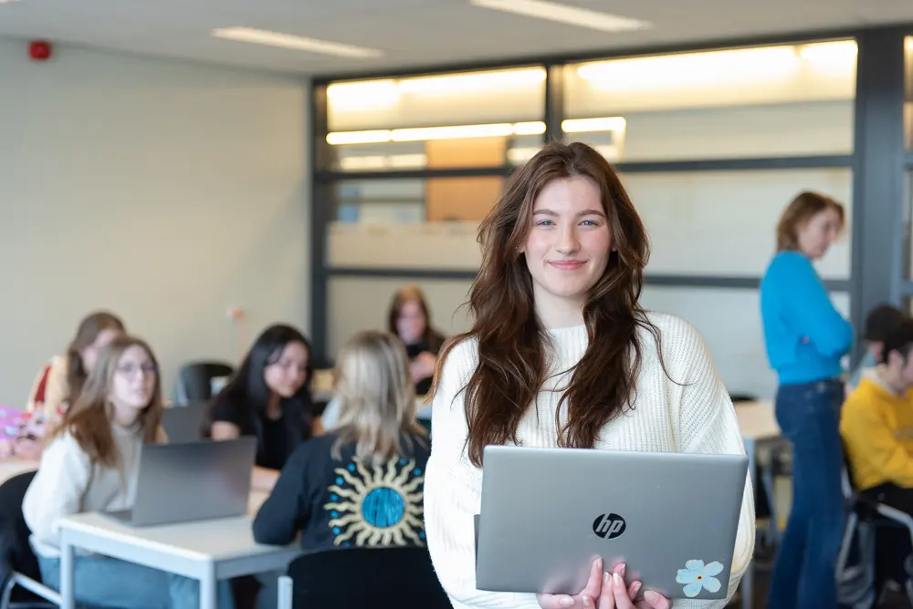 Klaslokaal met leerlingen waar op de voorgrond een leerling staat met een laptop in haar hand.