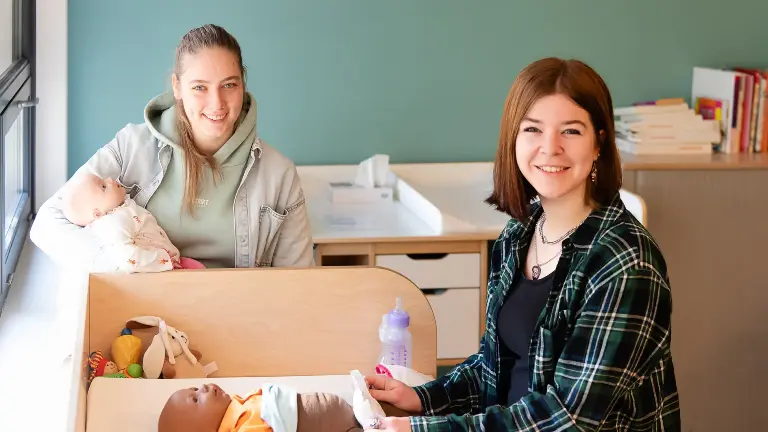 Twee personen in een kamer; één houdt een babypop vast, de ander is bezig met een babypop op een verschoontafel. Rondom liggen speelgoed, een babyfles en andere spullen.