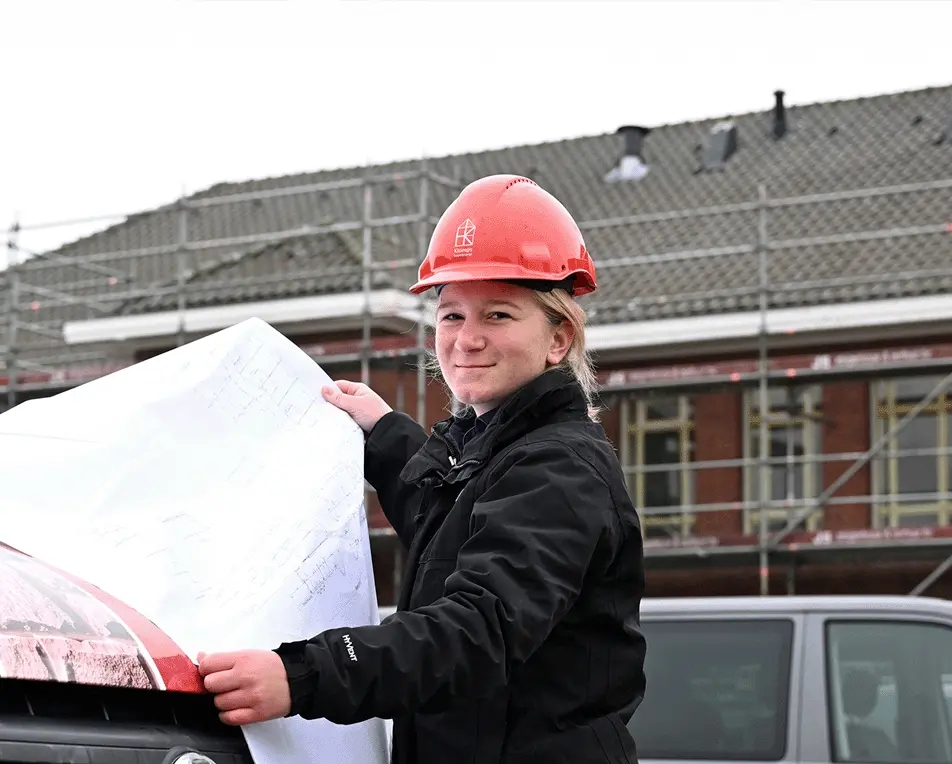Vrouw met rode veiligheidshelm heeft bouwtekeningen vast en staat buiten.