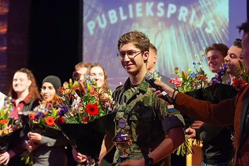 Groep mensen met bloemen, de middelst persoon houdt een trofee vast.