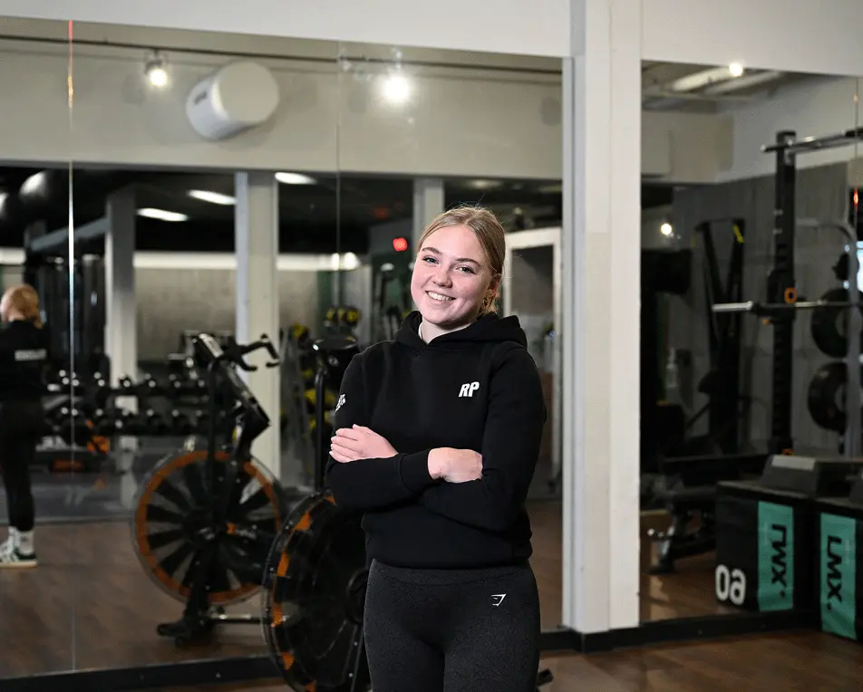 Vrouw in sportlegging en trui staat in een fitnessruimte.