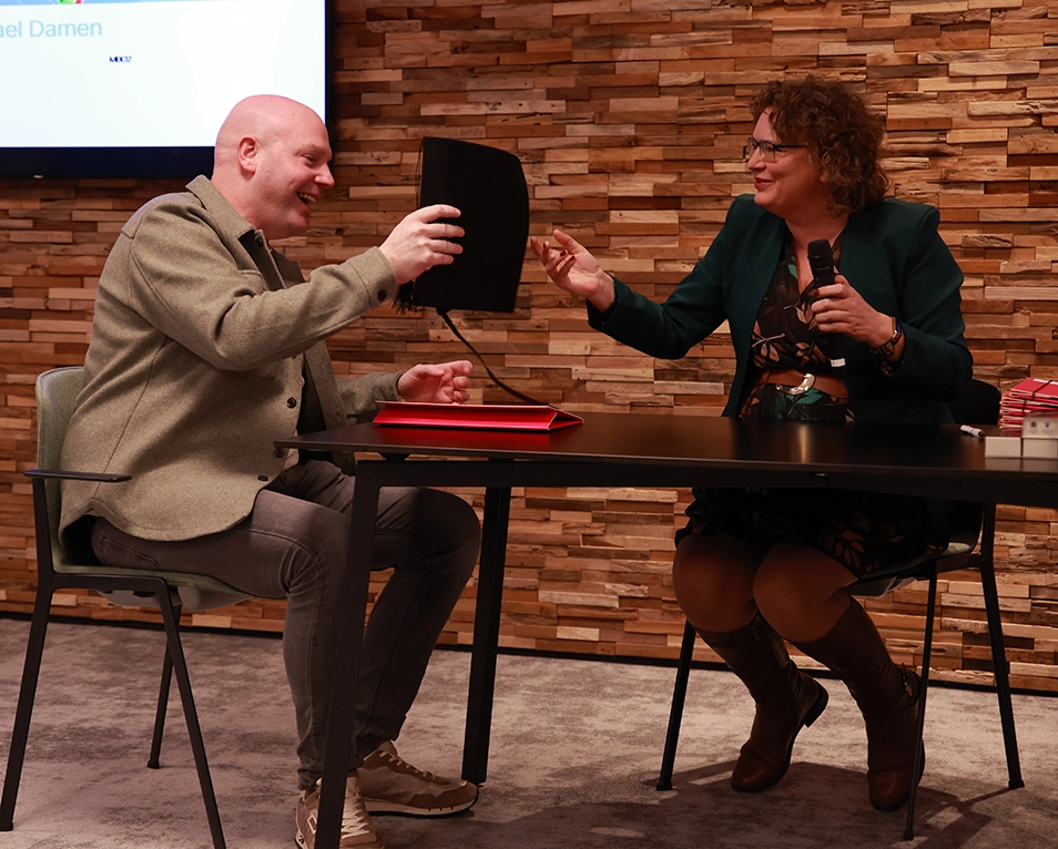 Michael Damen ontvangt zijn diploma en lacht tijdens de uitreiking, samen met een docent aan tafel.