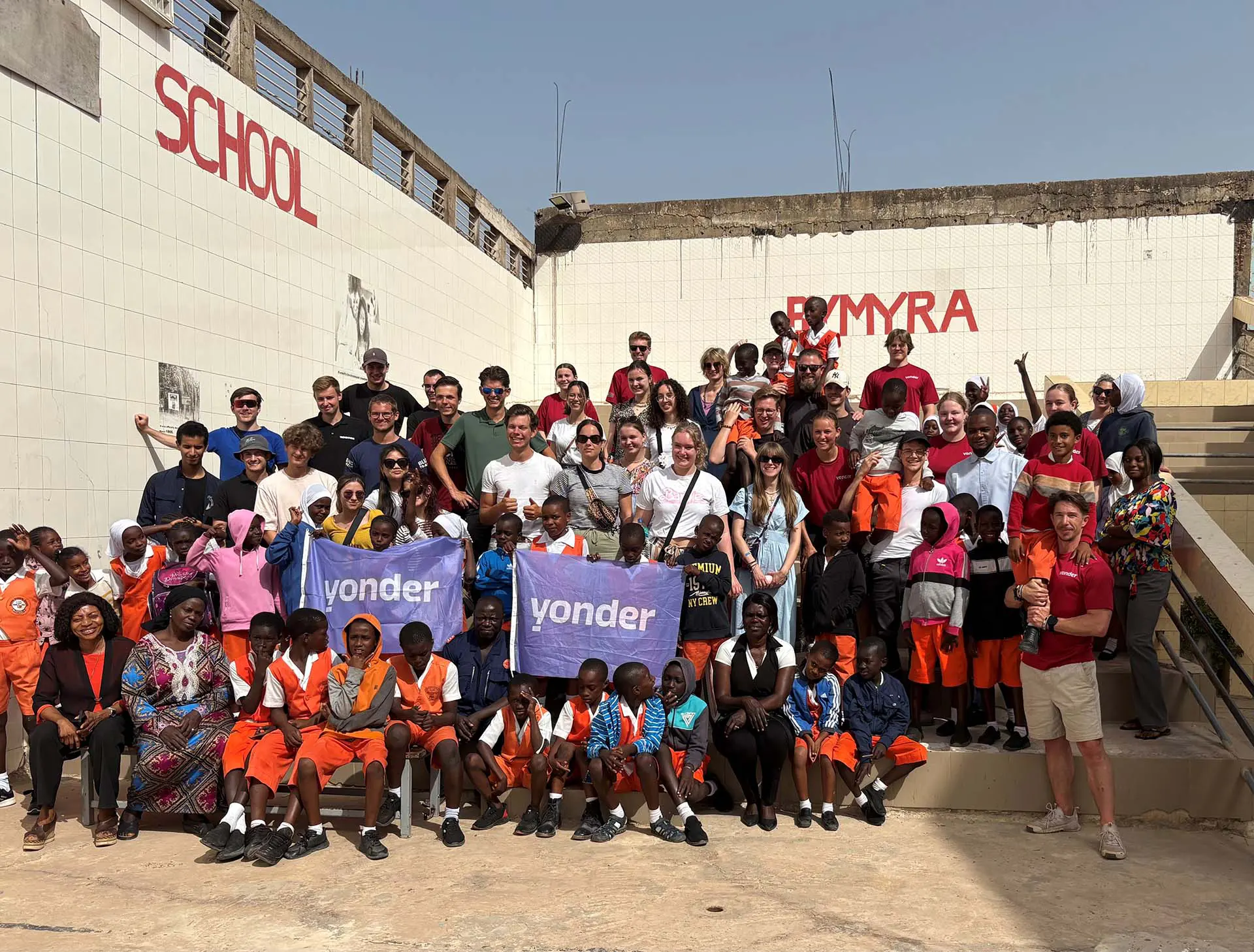 Grote groep mensen, waaronder kinderen in uniform en volwassenen, poseert buiten voor een witte betegelde muur.