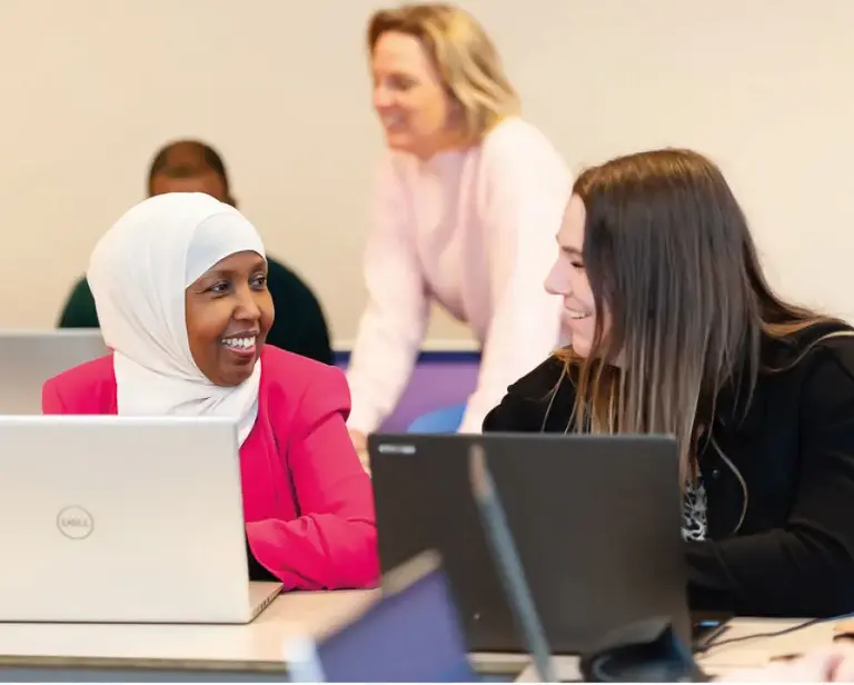 Een jonge dame met witte hoofddoek en een jonge dame met lang donker haar zitten lachend naast elkaar voor hun laptop.