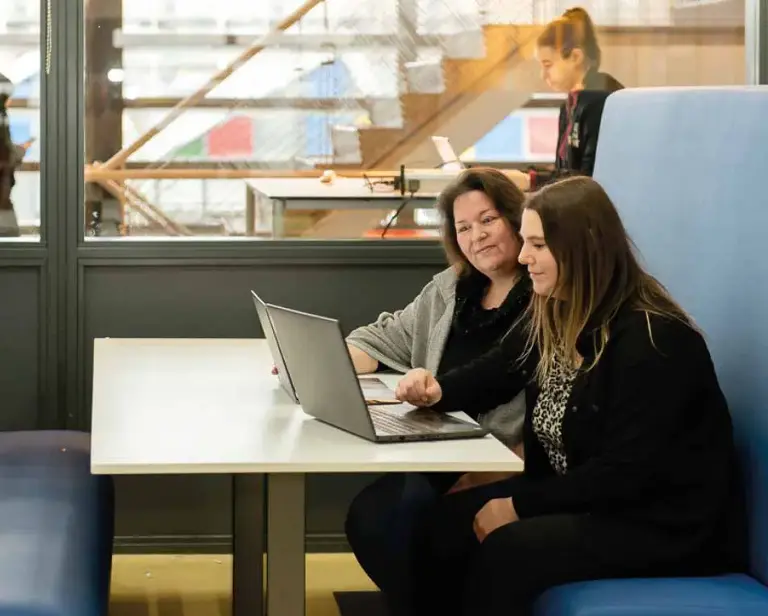 Een oudere en een jonge vrouw zitten op een blauwe bank en kijken naar hun laptop.