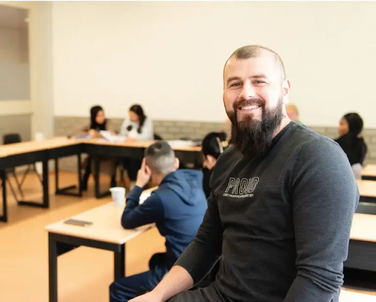 Man met lange baard en donker T-shirt zit in een klaslokaal, met leerlingen op de achtergrond.