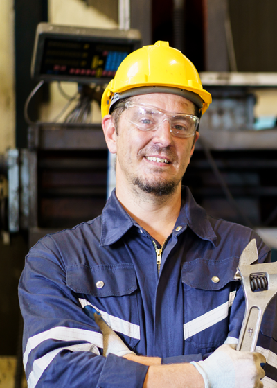 Man in donkerblauwe overall, met gele veiligheidshelm en veiligheidsbril, voor de achtergrond van een werkruimte.