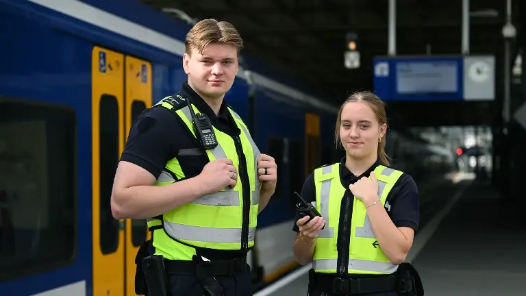Man en vrouw in gele hesjes staan op een perron, met een trein op de achtergrond.