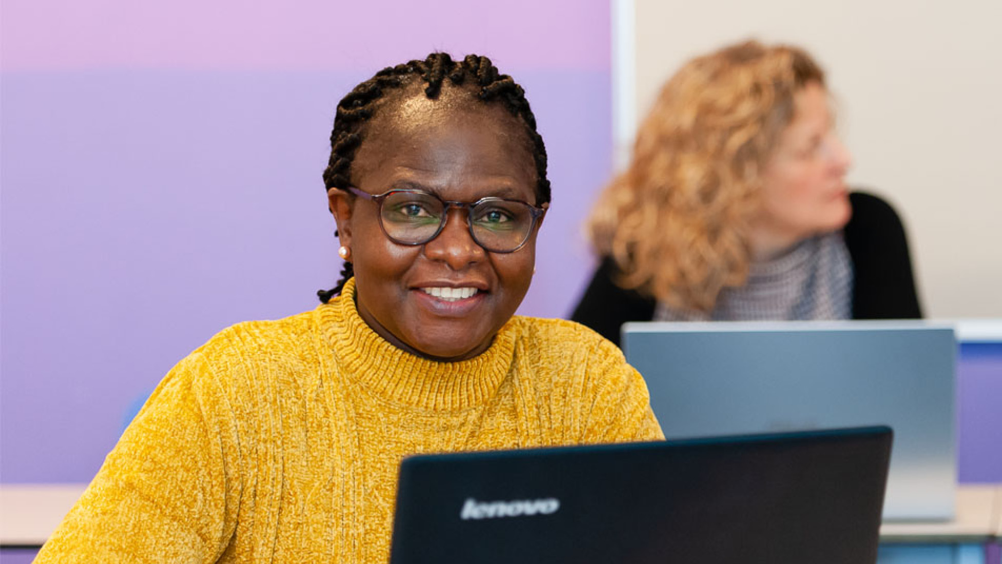 Vrouw in gele trui achter laptop in een klassikale omgeving.