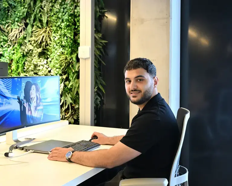 Man met baard zit in een kantoor achter een computer op een bureaustoel.