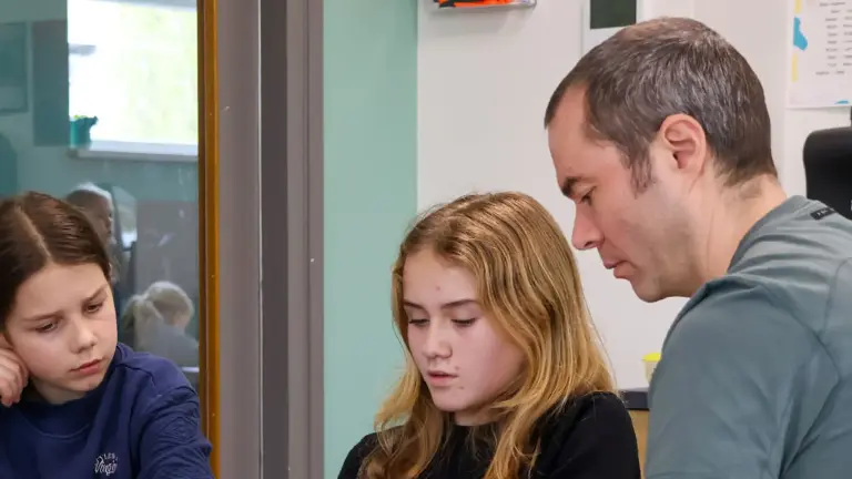 Man in grijs T-shirt geeft uitleg aan twee jongere meiden, die in een klassikale omgeving zitten.