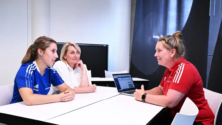 Drie dames, waarvan twee in sportoutfit en één in een witte blouse, hebben overleg aan een witte tafel.