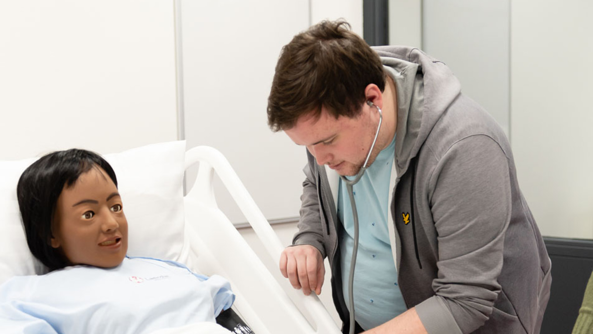 Man in grijs vest luistert met een stethoscoop naar de arm van een pop in een ziekenhuisbed.