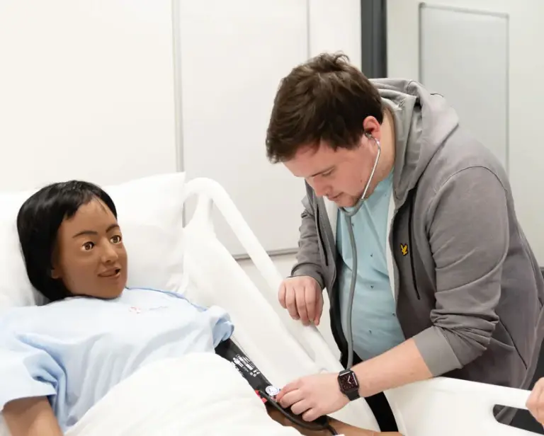 Man in grijs vest luistert met een stethoscoop naar de arm van een pop in een ziekenhuisbed.